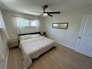 Un dormitorio con cama, mesita de noche, ventilador de techo, ventana con cortinas y puerta.