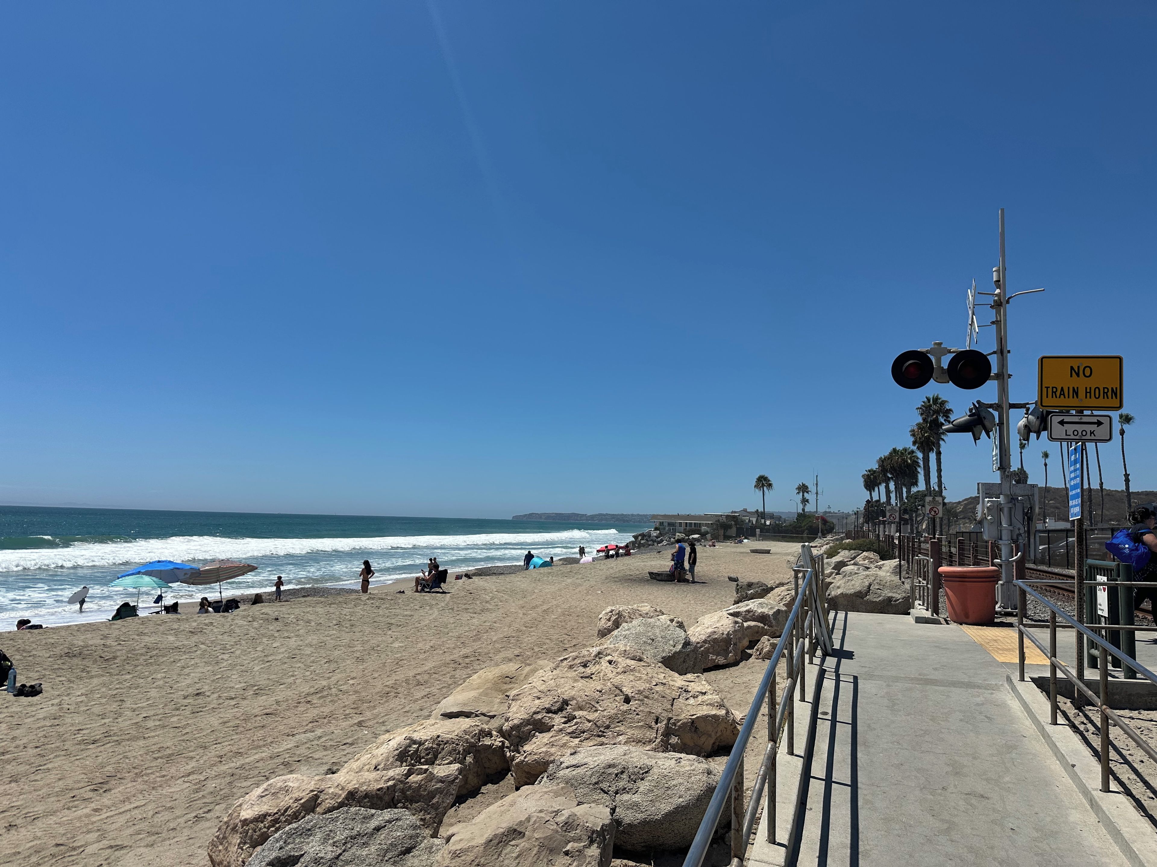 A beach with a concrete walkway, a 'NO TRAIN HORN' sign, and palm trees in the background.
