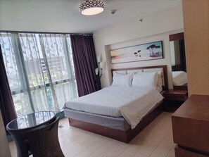 A hotel room with a bed, a chair, a painting, and a view through the window. - One Uptown Residence 30H (Taguig City)