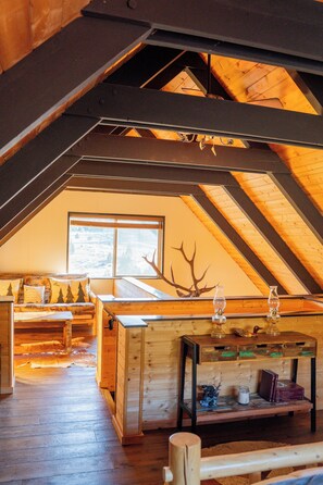 Ein gemütliches Dachzimmer mit geneigter Decke, Holzbalken, einem großen Fenster, einer Couch, einem Tisch und einer Dekoration aus Hirschgeweihen.