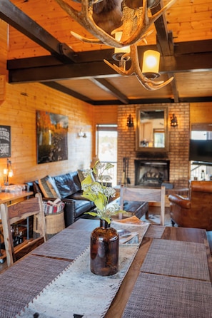 A cozy interior with a fireplace, wooden walls, and a dining area with a vase of flowers. - Riverfront Cabin on 3 Acres Fire pit + Mountain Views (Hatch)