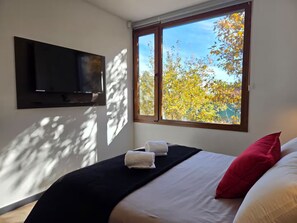 Een slaapkamer met een bed, een flatscreen televisie aan de muur en een raam met uitzicht op bomen.