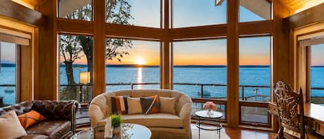 A spacious room with a large window overlooking a body of water at sunset, furnished with leather sofas and a glass coffee table.