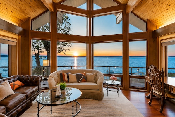 A spacious room with a large window overlooking a body of water at sunset, furnished with leather sofas and a glass coffee table.