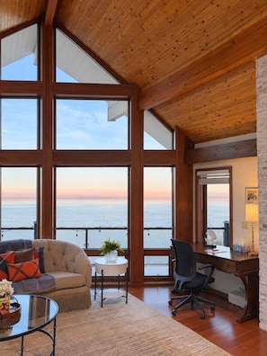 A spacious room with large windows, a wooden ceiling, a cozy seating area, and a desk with a lamp.