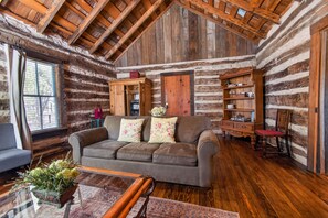 A log cabin interior with a wooden ceiling, a sofa with floral cushions, a wooden cabinet, and a bookshelf. - Historic Cabin w/King Bed + Walk to Wineries! (Fredericksburg)