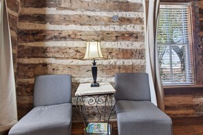 A room with a rustic wooden wall, a small table with a lamp, two grey armchairs, and a window with blinds. - Historic Cabin w/King Bed + Walk to Wineries! (Fredericksburg)