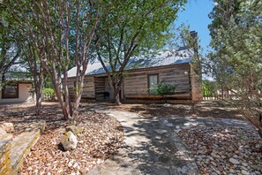 A rustic log cabin with a stone foundation, surrounded by a gravel path and trees. - Historic Cabin w/King Bed + Walk to Wineries! (Fredericksburg)