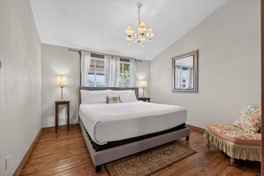 A bedroom with a large bed, two bedside lamps, a chandelier, a mirror, and a floral chair. - Historic Cabin w/King Bed + Walk to Wineries! (Fredericksburg)