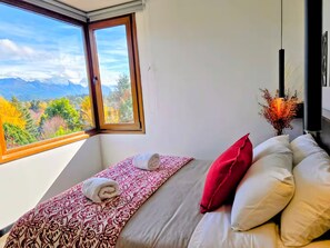 Ein Schlafzimmer mit großem Fenster, einem Bett mit gemusterten Betttextilien, einem roten Kissen und einer Vase mit Trockenblumen.