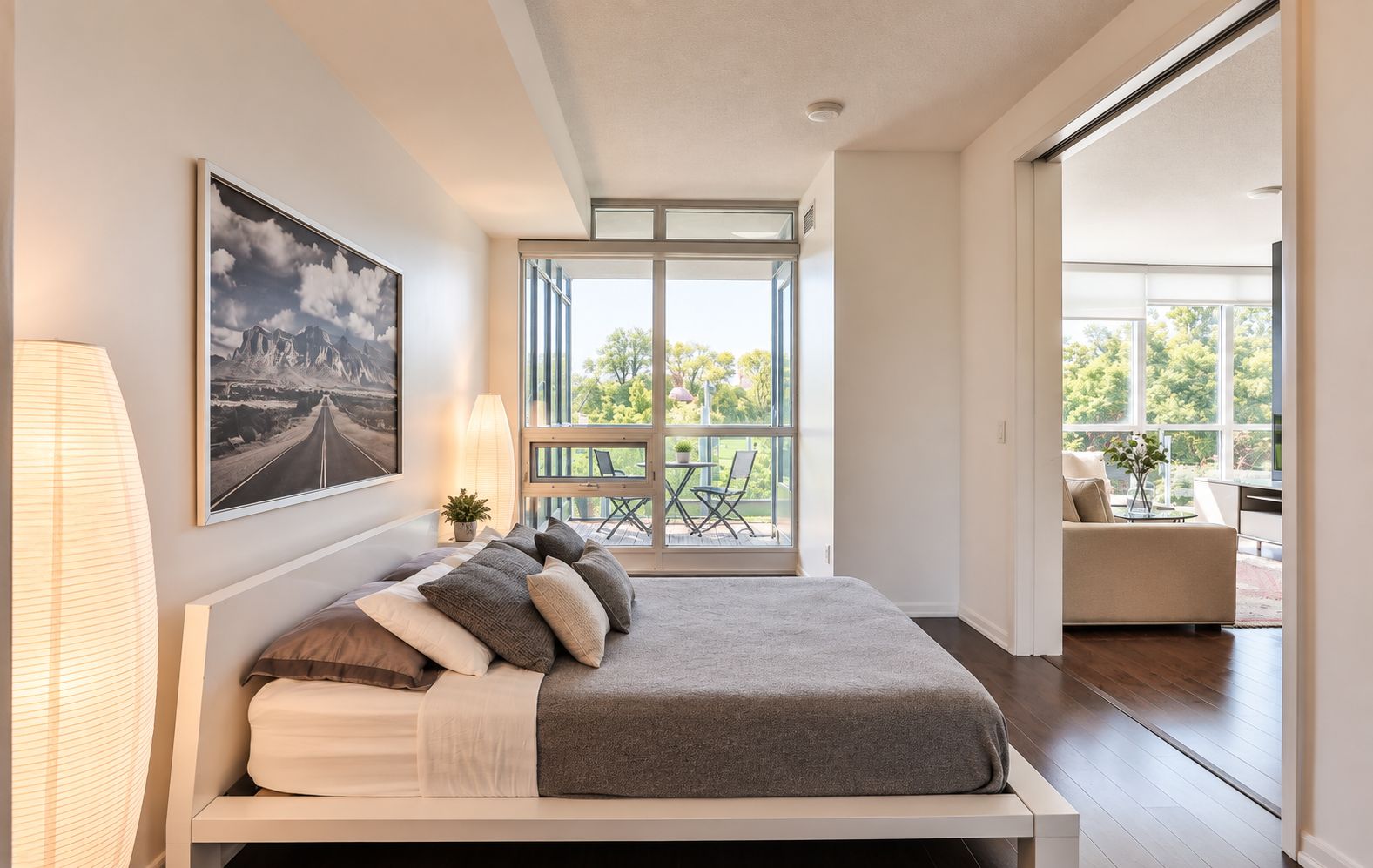 A modern bedroom with a large bed, a view of the outdoors through a sliding glass door, and a painting on the wall.