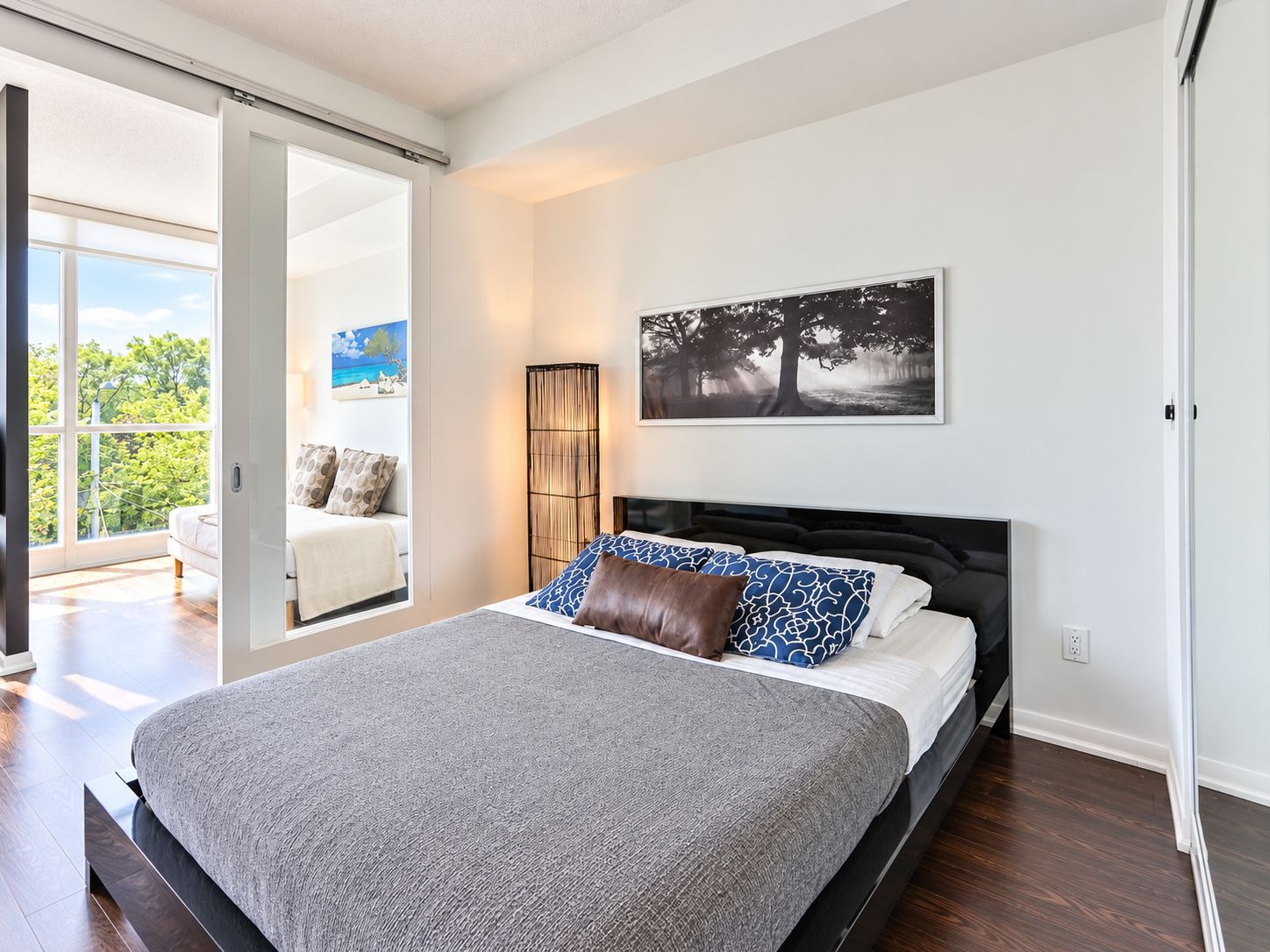 A bedroom with a large bed, a view of the outdoors through a sliding door, and a painting on the wall.