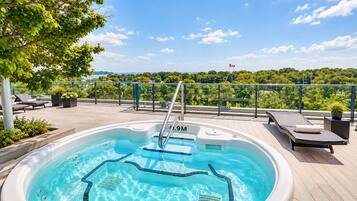 A hot tub with a view of a forest and a flag, surrounded by lounge chairs and potted plants.