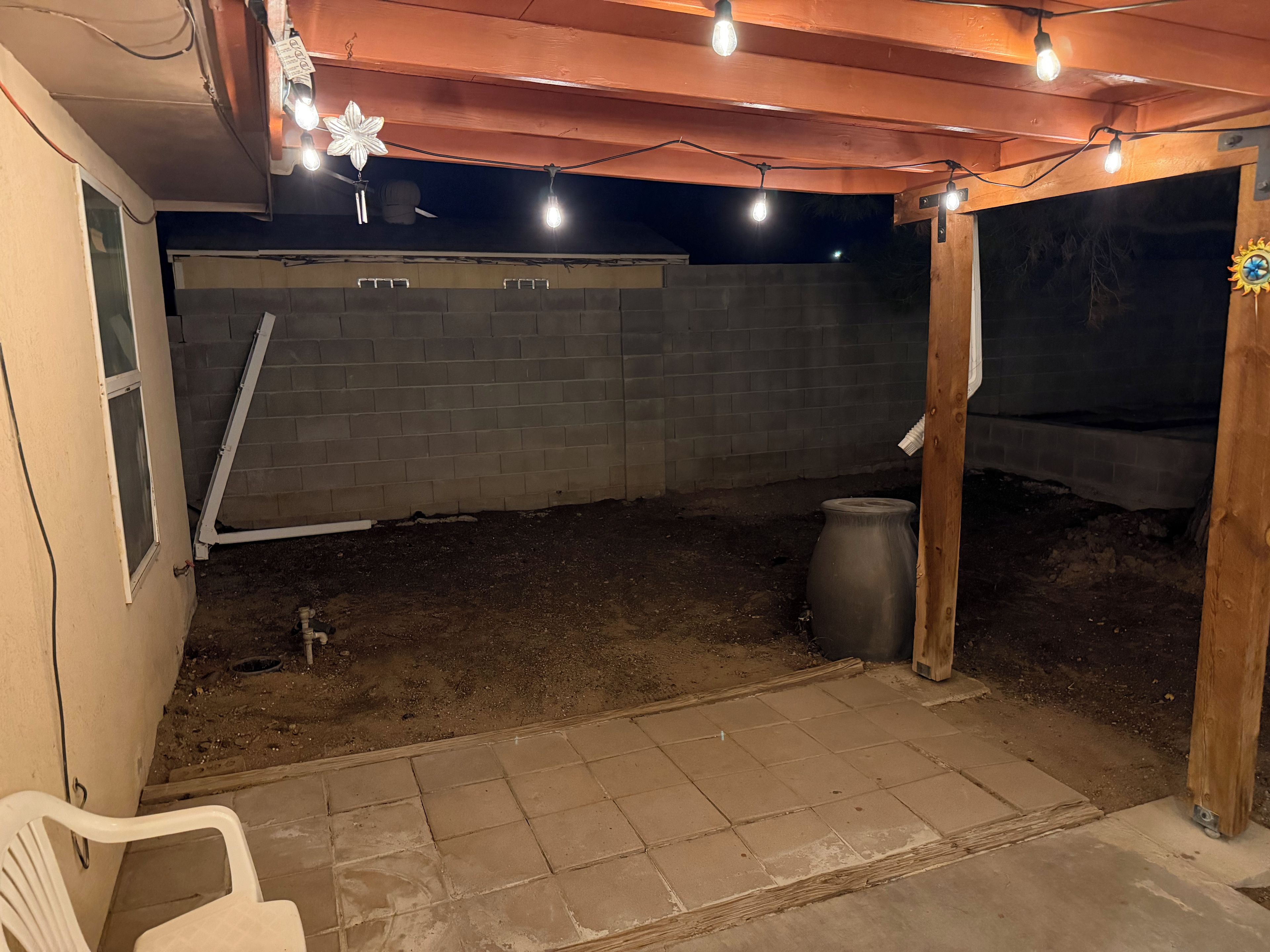 Un espace extérieur abrité, doté d’un mur en briques, d’une pergola en bois illuminée de guirlandes lumineuses et d’une chaise en plastique blanche.