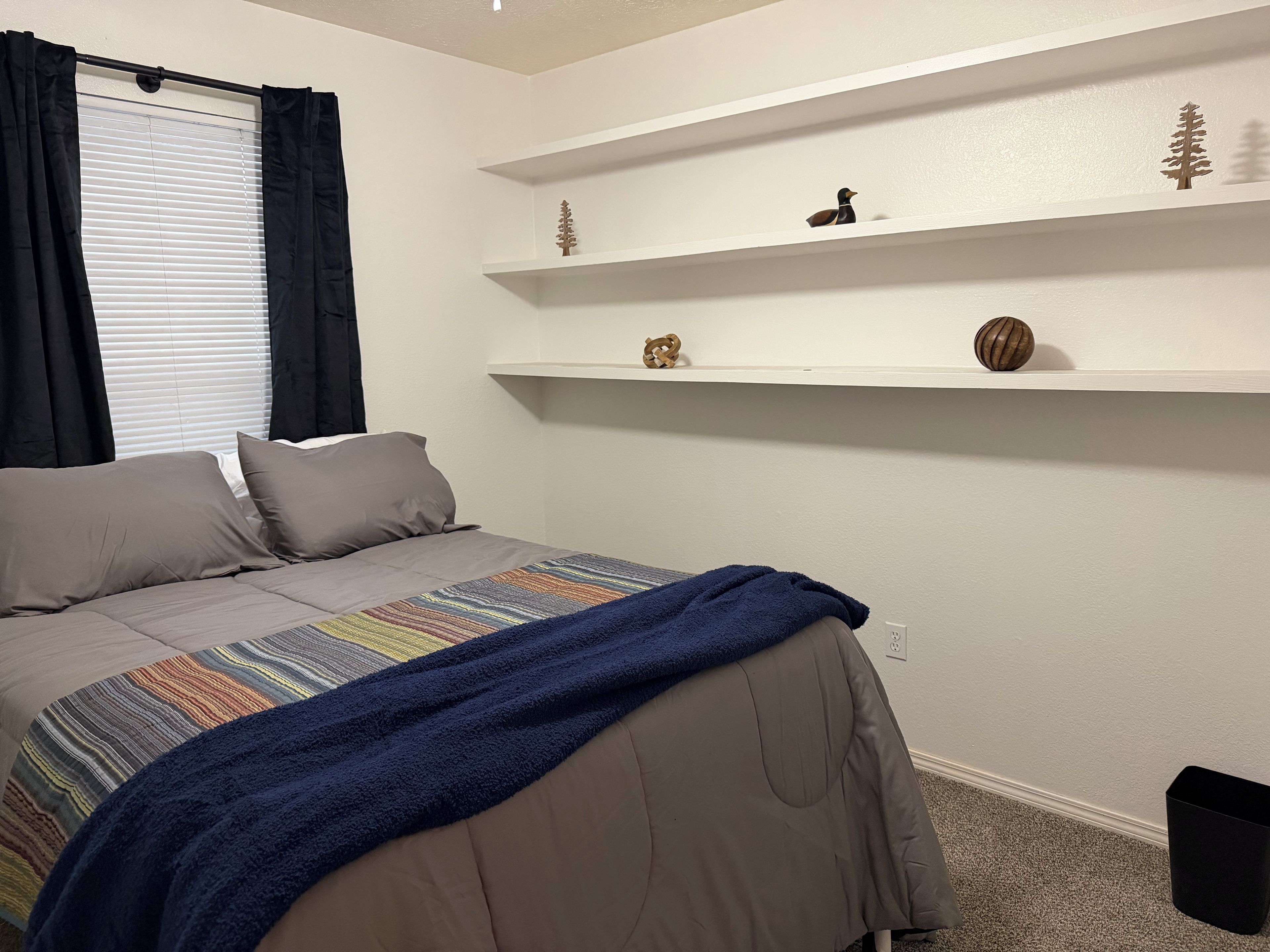 Une chambre à coucher avec un lit, des oreillers, une couverture, une fenêtre avec des rideaux et des étagères ornées d’objets décoratifs.