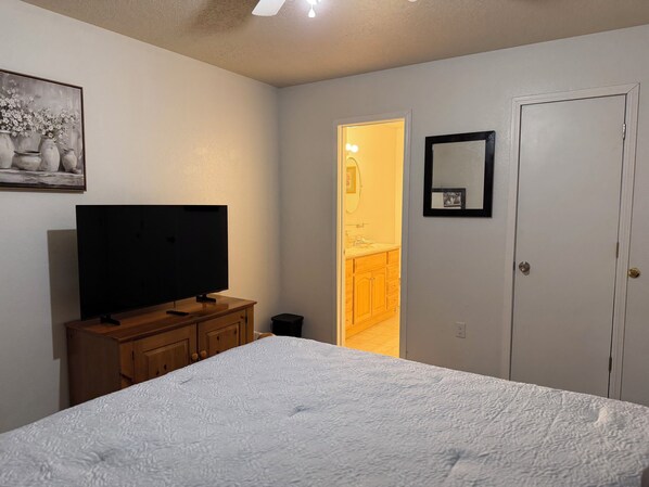 Une chambre à coucher avec un lit, un téléviseur posé sur une commode en bois, une porte donnant sur une salle de bain et un tableau encadré accroché au mur.
