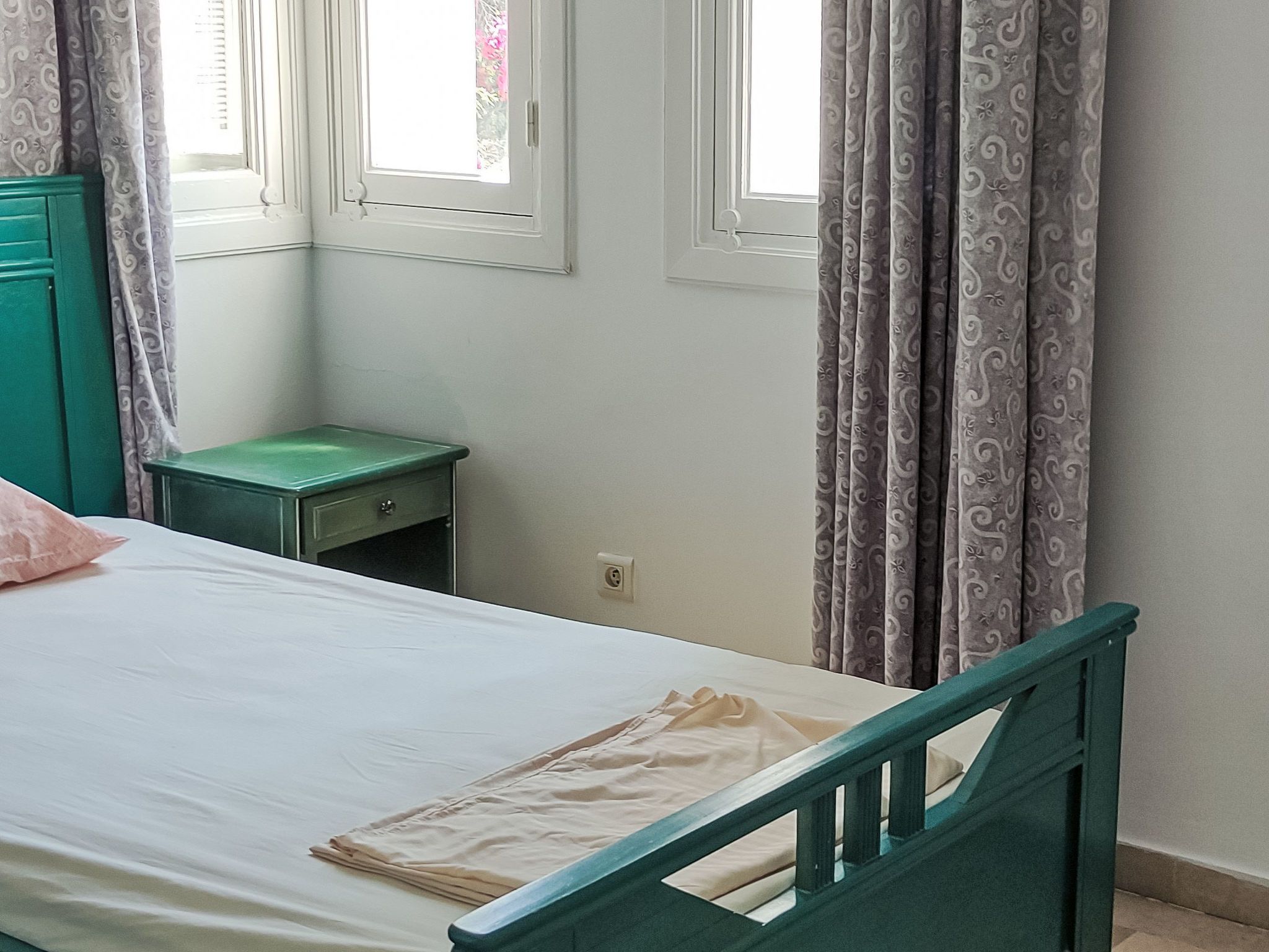 Une chambre avec un lit simple, une table de chevet, une armoire verte et des rideaux à motifs.