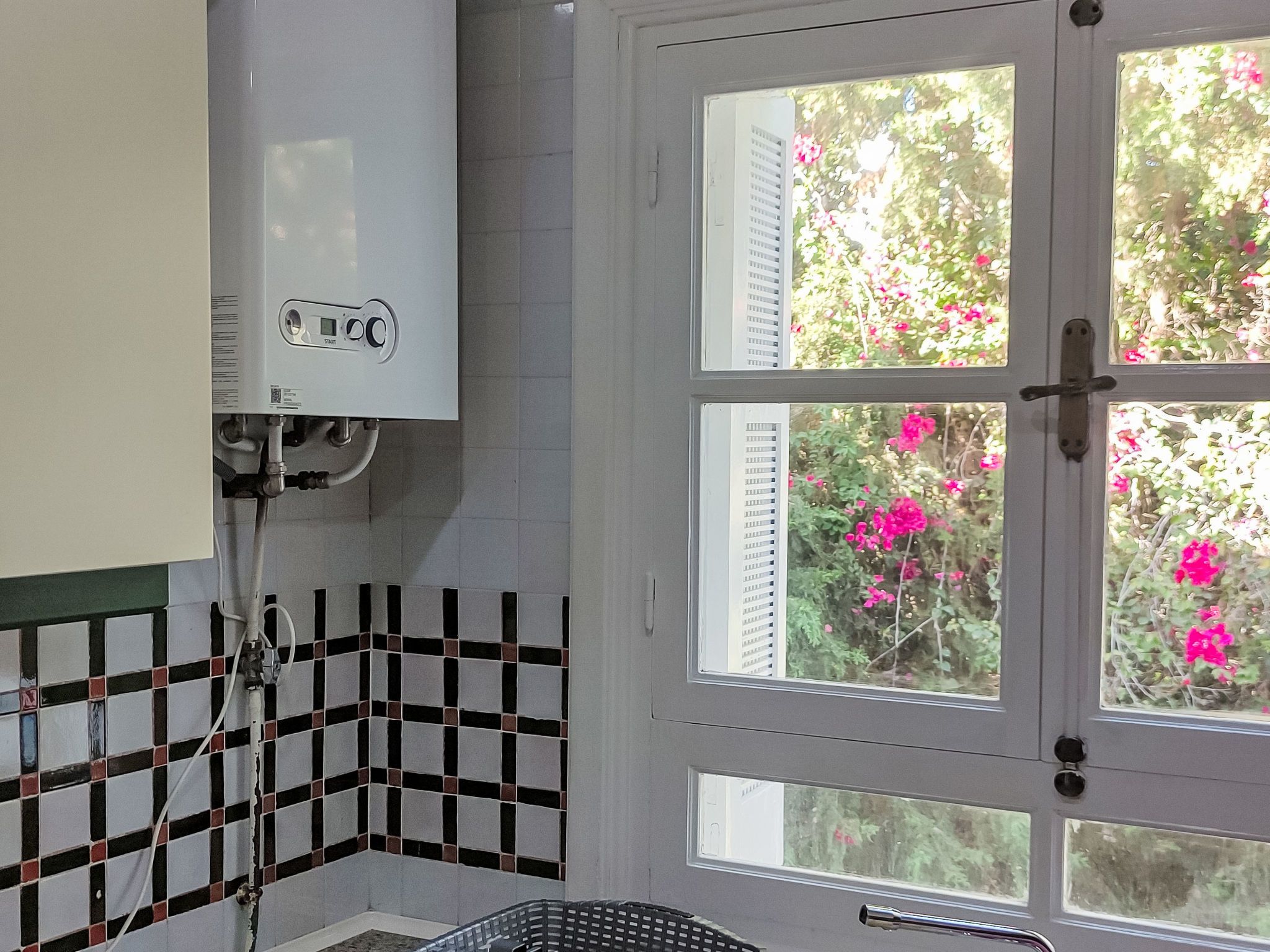 A bathroom with a white tiled wall, a shower area, and a window with a view of pink flowers.
