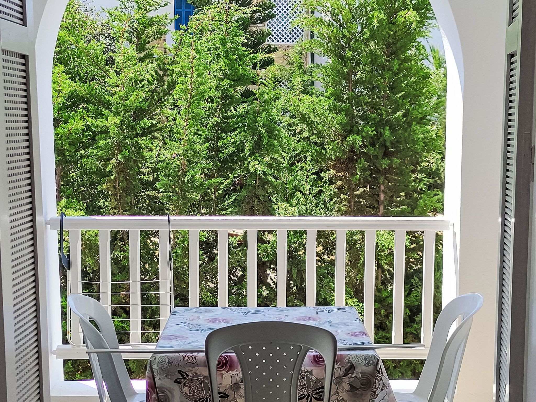 Ein Balkon mit Tisch und Stühlen, mit Blick auf einen Garten mit Bäumen und einem blauen Gebäude im Hintergrund.