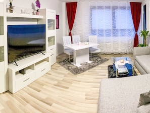 Ein modernes Wohnzimmer mit einem Flachbildfernseher, einem weißen Sofa, einem Esstisch mit Stühlen und einem großen Fenster mit roten Vorhängen.