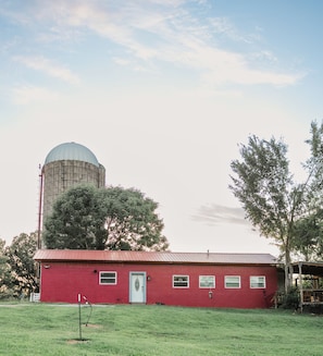 A red barn with a white door and windows, a silo in the background, and a grassy field.