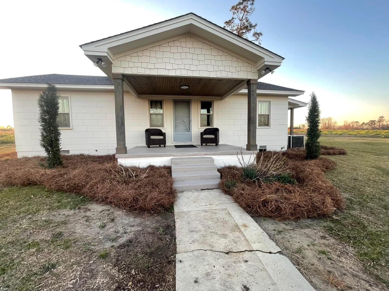 A single-story house with a front porch, two chairs, and a small tree.