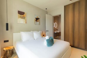 Une chambre d’hôtel avec un grand lit, une table de chevet en bois, deux interrupteurs d’éclairage fixés au mur, un tableau et une salle de bain visible à travers une porte ouverte.