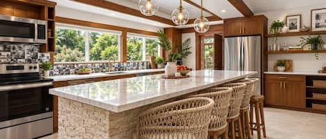 A modern kitchen with a large island, bar stools, stainless steel appliances, and a view of greenery outside.
