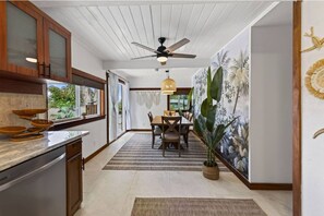 A kitchen with a dining area, a ceiling fan, and a wall with tropical mural. - Kona Gathering Home | Sunset Views, Lanai & BBQ (Kailua-Kona)