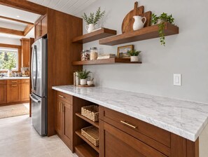 A kitchen with wooden cabinets, a marble countertop, and built-in shelves with decorative items. - Kona Gathering Home | Sunset Views, Lanai & BBQ (Kailua-Kona)