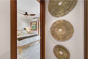 A bedroom with a bed, a ceiling fan, and three wall-mounted decorative items. - Kona Gathering Home | Sunset Views, Lanai & BBQ (Kailua-Kona)