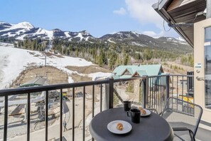 Ein Balkon mit Tisch, einem Teller mit Gebäck und einer Tasse, mit Blick auf eine Berglandschaft und einem Skiort im Hintergrund.