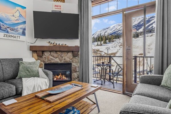 Ein gemütliches Wohnzimmer mit Kamin, Flachbildfernseher und Blick auf eine verschneite Berglandschaft durch eine Glastür.