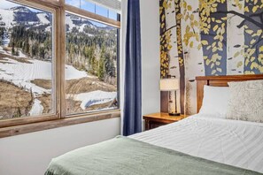 Ein Hotelzimmer mit großem Fenster und Blick auf eine verschneite Landschaft, einem Bett mit weißer Bettwäsche, einem Nachttisch aus Holz mit Lampe sowie einer Wand mit einem Wandgemälde zum Thema Natur.