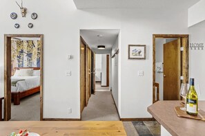 Quarto de hotel com uma cama, uma mesa de madeira com uma garrafa de vinho e um corredor que leva a outros quartos.