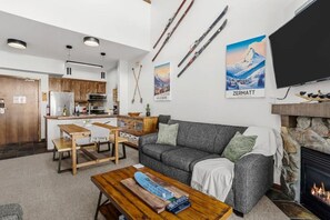 Sala de estar con un sofá gris, una mesa de centro de madera y una chimenea. Hay una zona de cocina con armarios de madera y un refrigerador. Equipo de esquí exhibido en la pared.