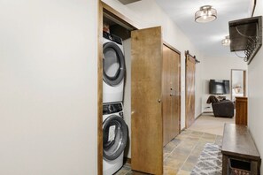 A laundry room with a washing machine and dryer, a wooden door, a television, and a seating area with a bench. - Golden Alpine Escape 3BR+Den HotTub+Ski In/Out (Golden)