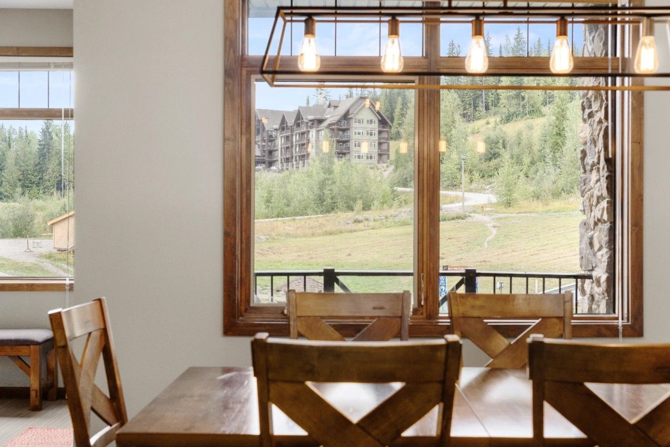 A dining area with a table and chairs, a large window with a view of a building and trees, and a hanging light fixture.