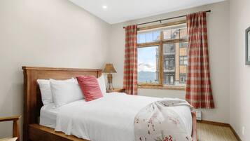 A bedroom with a wooden bed, white bedding, a red pillow, a lamp, and a window with plaid curtains.