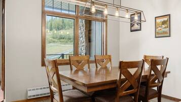 Un espace repas avec une table et des chaises en bois, de grandes fenêtres donnant sur un paysage verdoyant et un luminaire suspendu.