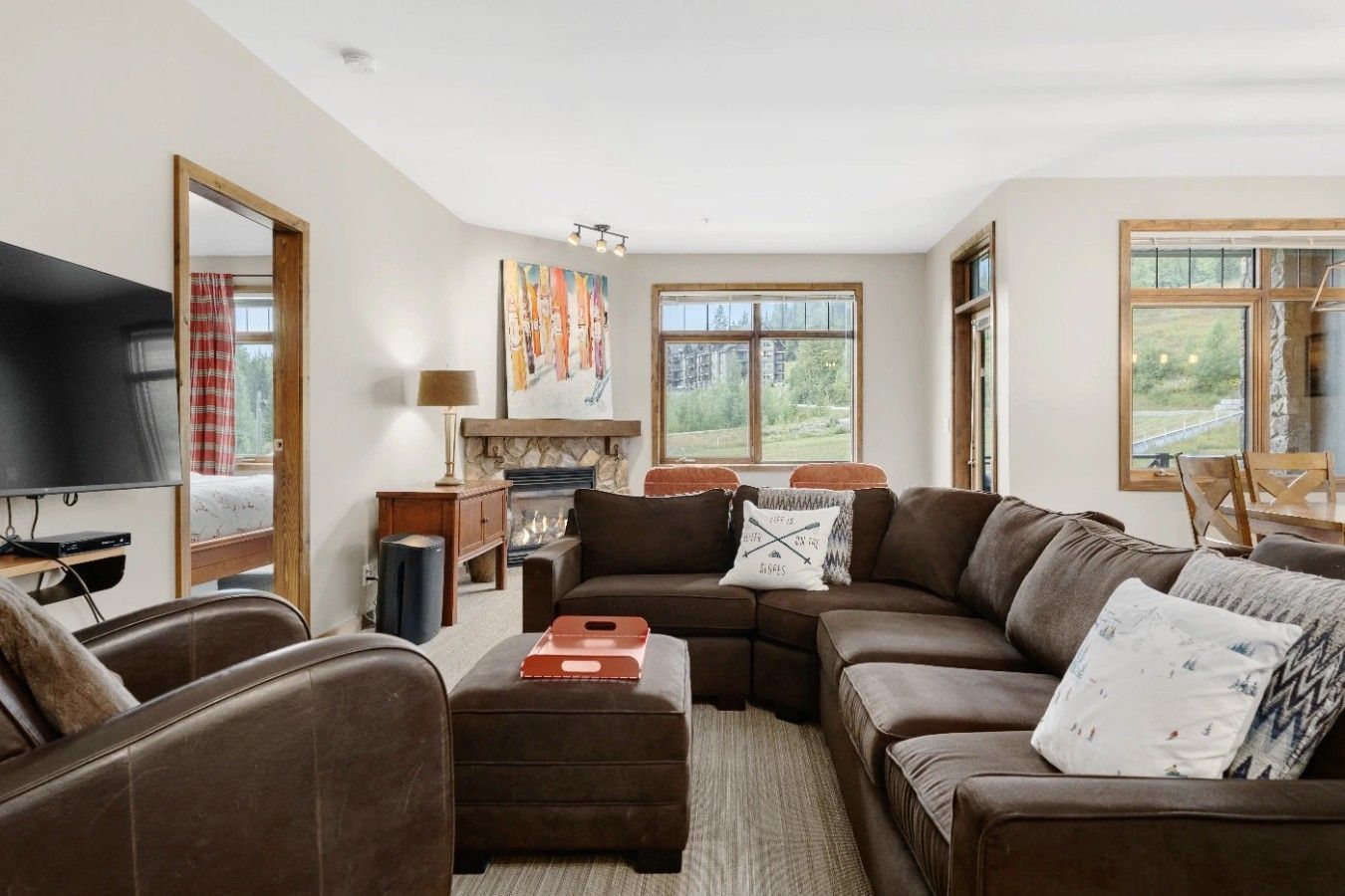 A living room with a large brown sofa, a television, a fireplace, and a view of the outdoors through large windows.