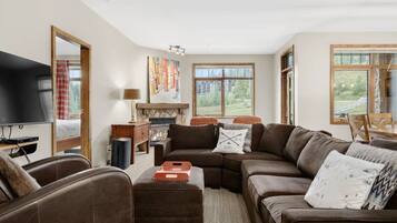 A living room with a large brown sofa, a television, a fireplace, and a view of the outdoors through large windows.