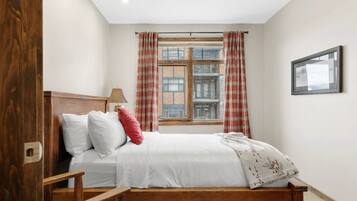 A bedroom with a wooden bed, a window with curtains, a lamp, and a framed picture on the wall.