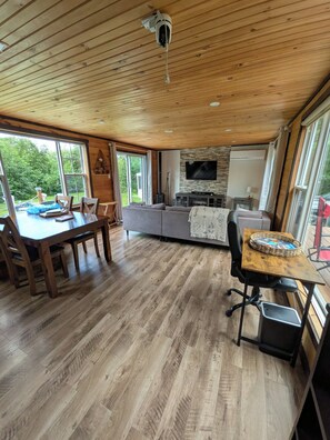A spacious living area with a wooden ceiling, a dining table with chairs, a gray sofa, and a television mounted on the wall. - Short walking distance to beach. With fire pit, BBQ,n pedal boat. (Beaubassin-est)