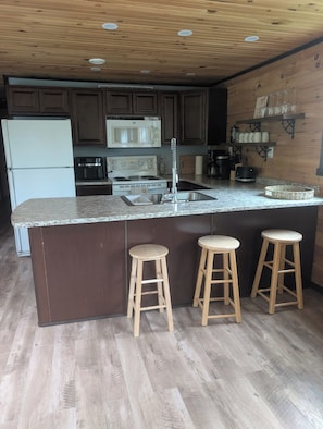 A kitchen with a central island, wooden stools, a refrigerator, a microwave, a stove, and a coffee maker. - Short walking distance to beach. With fire pit, BBQ,n pedal boat. (Beaubassin-est)
