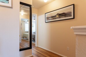 Un couloir doté d’un plancher en bois, une peinture de paysage encadrée accrochée au mur, et une porte donnant sur une chambre à coucher équipée d’un lit et d’un ventilateur de plafond.