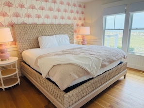 A bedroom with a large bed, two bedside tables with lamps, a patterned wall, and a window with a view. - Ocean Views & Southern Charm on Bald Head Island (Bald Head Island)