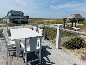 Une terrasse en bois avec une table et des chaises blanches, donnant sur une plage et des maisons.