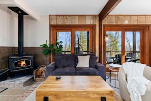 A cozy living room with a fireplace, a sofa, a coffee table, and a view through large windows. - Blackrock Canopy (Nellysford)