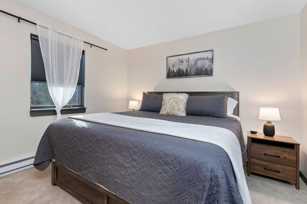 A bedroom with a bed, bedside tables, a window with sheer curtains, and a wall-mounted painting.
